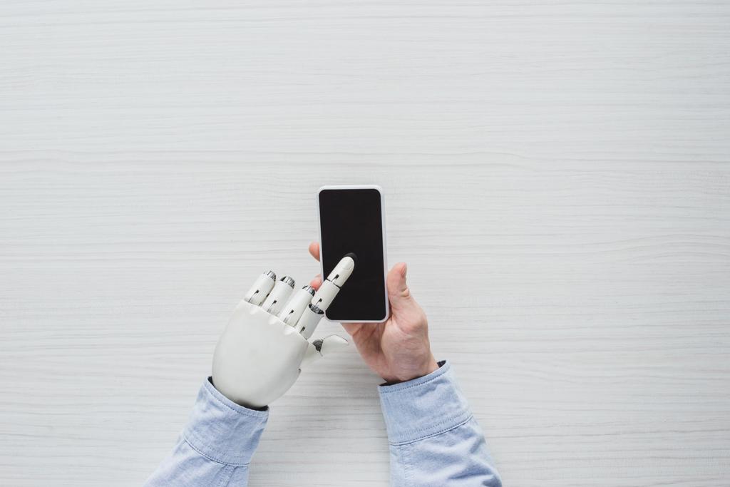 περικομμένη εικόνα του ανθρώπου με το cyborg χέρι χρησιμοποιώντας smartphone με κενή οθόνη πάνω από το ξύλινο τραπέζι  - Φωτογραφία, εικόνα