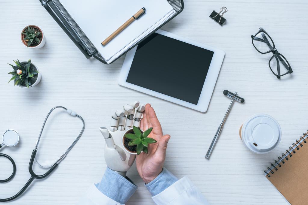 περικομμένη εικόνα αρσενικό ο γιατρός με προσθετική βραχίονα κατέχουν γλάστρες φυτών στο τραπέζι με ιατρικά εργαλεία και ψηφιακή δισκίο  - Φωτογραφία, εικόνα