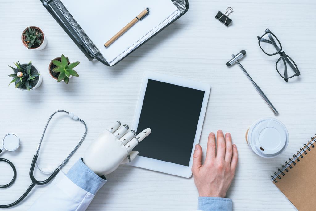 обрезанное изображение врача-мужчины с протезной рукой с помощью цифрового планшета с чистым экраном за столом с медицинскими инструментами
  - Фото, изображение