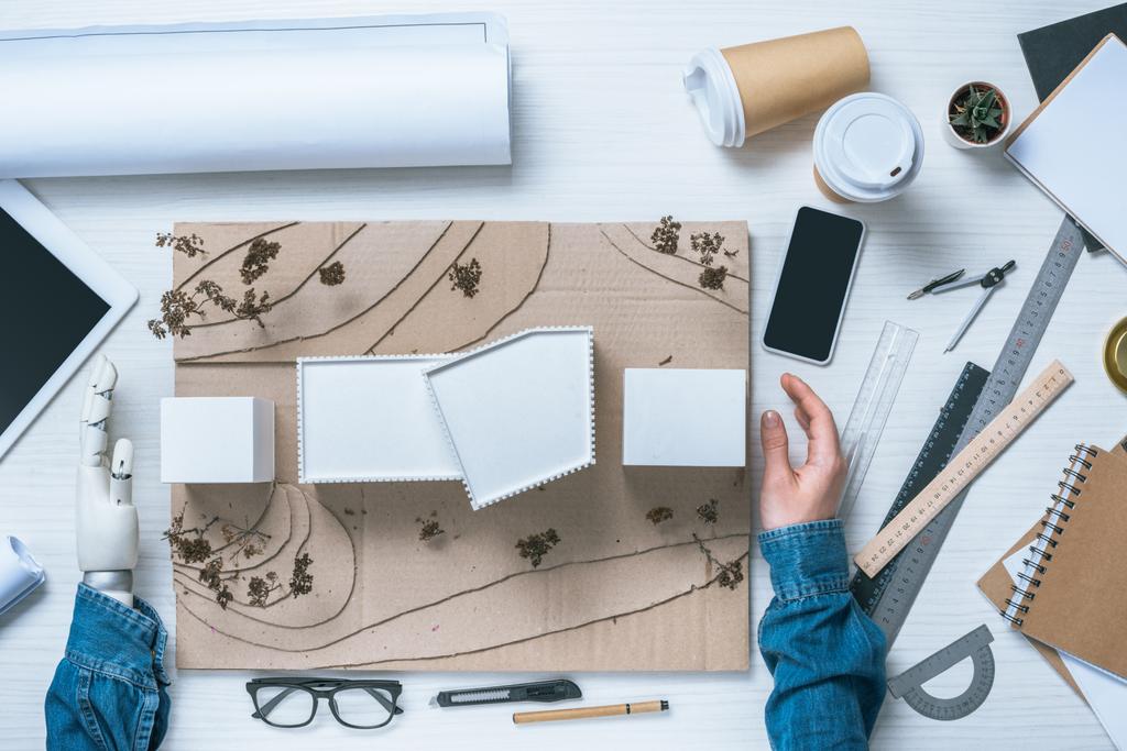 περικομμένη εικόνα του αρσενικό αρχιτέκτονα με προσθετική βραχίονα κάνοντας μοντέλο του οίκου στο τραπέζι με φλιτζάνια καφέ, κυβερνήτες και σχεδιάγραμμα  - Φωτογραφία, εικόνα