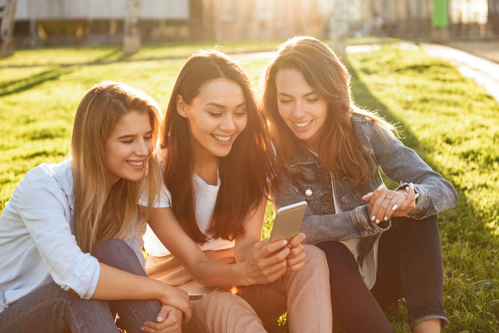 Kuva onnellinen kolme ystävää naiset istuvat nurmella puistossa ulkona etsivät sivuun matkapuhelimella
. - Valokuva, kuva