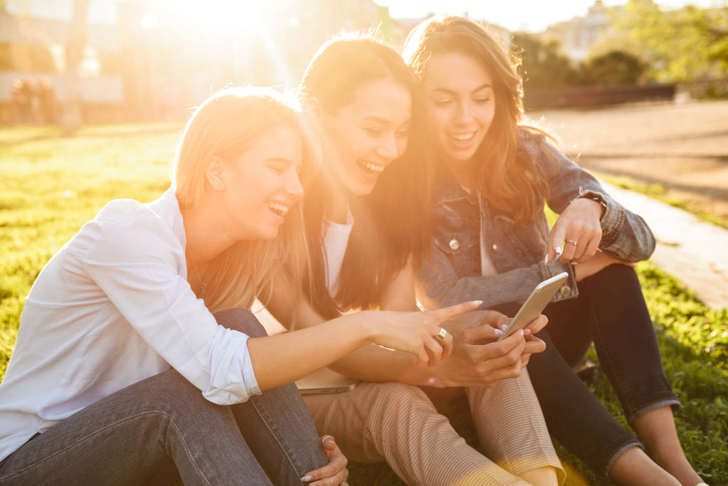 Photo de heureux trois amis femmes assises sur l'herbe dans le parc à l'extérieur regardant de côté en utilisant un téléphone mobile
. - Photo, image