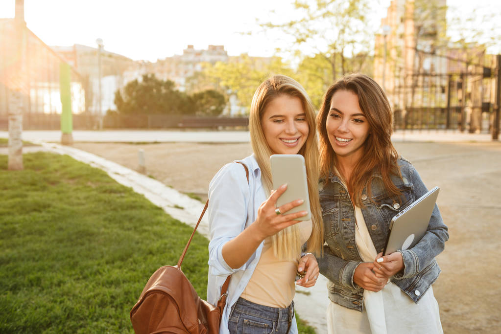 Açık havada tutarak dizüstü bilgisayar ve cep telefonuyla birbirleriyle konuşurken Park mutlu iki arkadaş kadınların görüntü. - Fotoğraf, Görsel