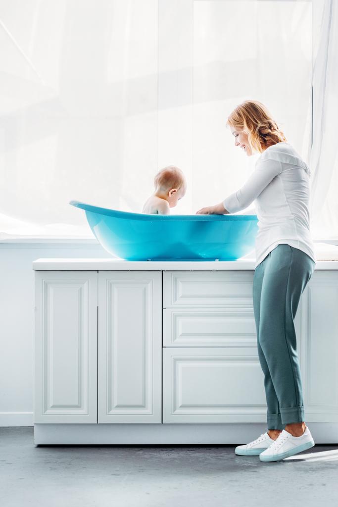 vista lateral da mãe banho criança na banheira de plástico em casa
 - Foto, Imagem