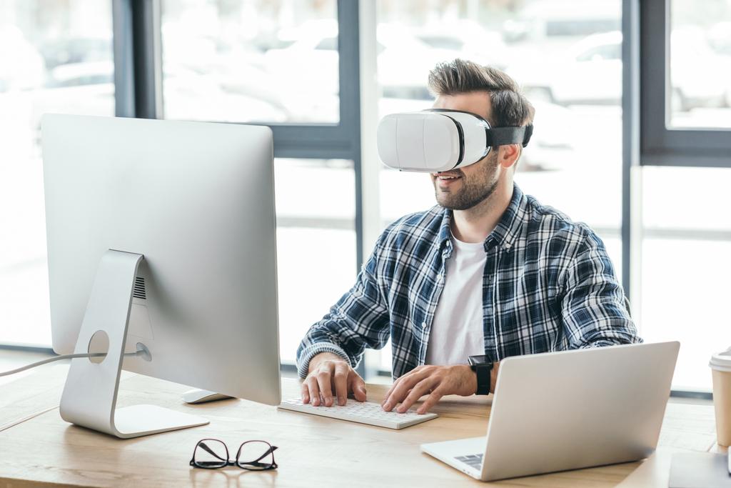 молодий чоловік у гарнітурі віртуальної реальності використовує настільний комп'ютер та ноутбук на робочому місці
   - Фото, зображення
