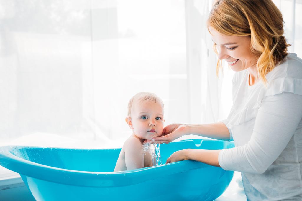 Seitenansicht einer glücklichen Mutter, die ihr entzückendes kleines Kind zu Hause in einer Plastikbadewanne wäscht - Foto, Bild