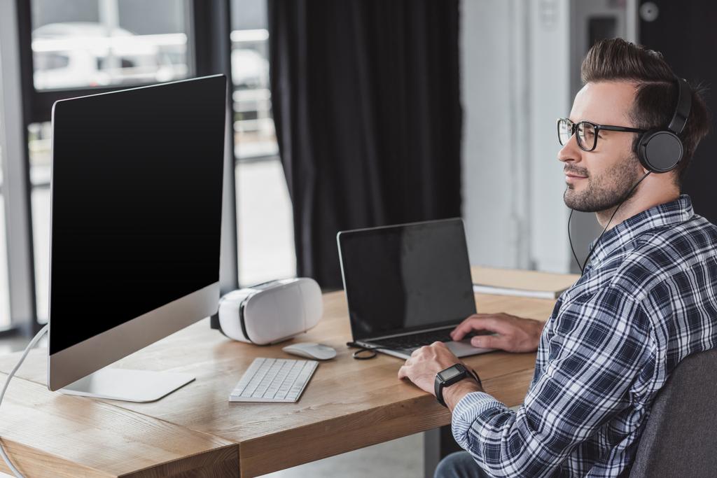 przystojny uśmiechający się młody programista w słuchawki i okulary za pomocą laptopa i komputera stacjonarnego - Zdjęcie, obraz