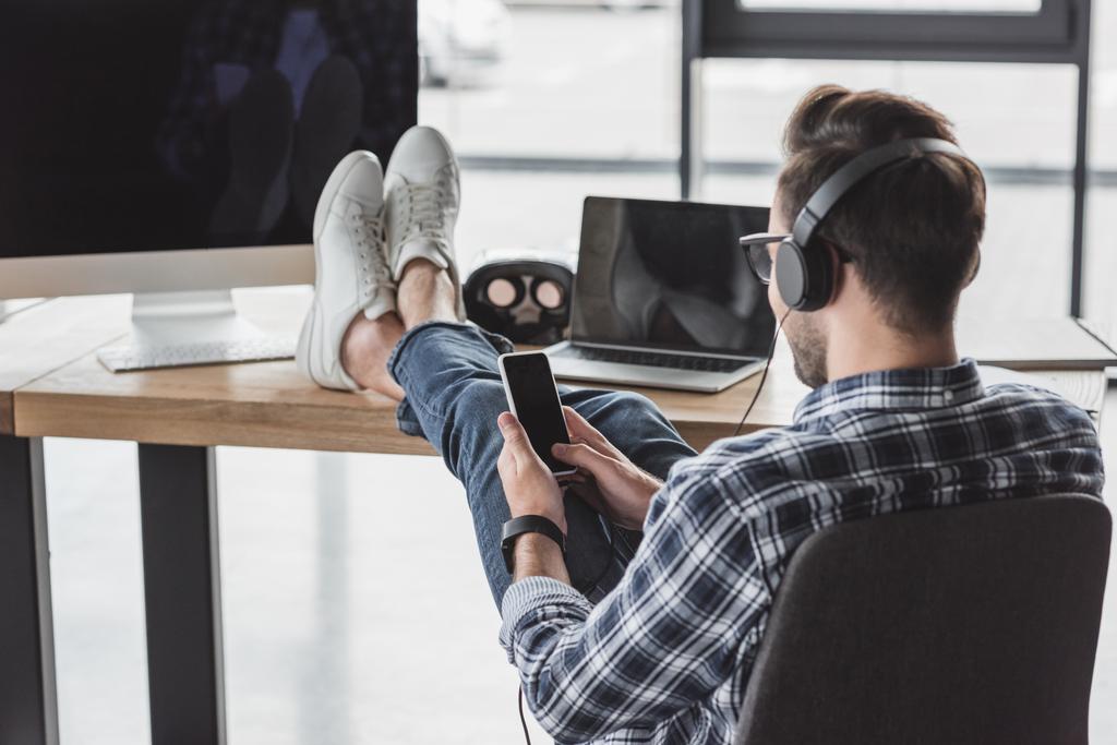 junger Mann mit Kopfhörer mit Smartphone, während er mit Beinen auf dem Tisch mit Laptop und Desktop-Computer sitzt - Foto, Bild