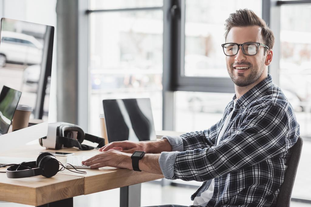 csinos, fiatal férfi a kamera munka közben a asztali számítógép és laptop mosolyogva szemüvegek - Fotó, kép