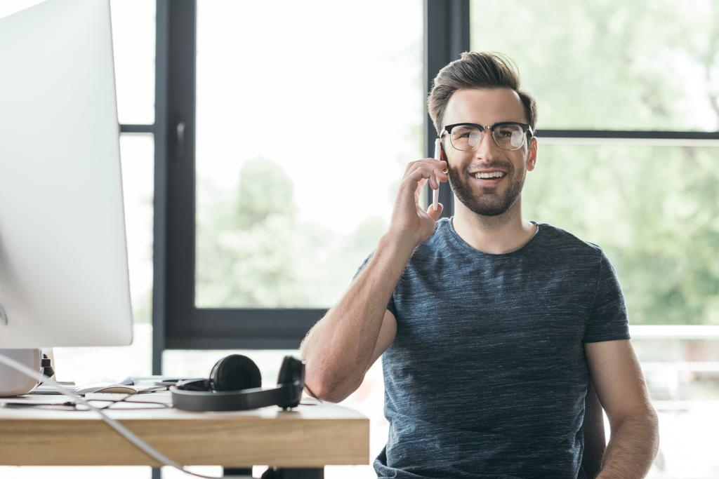 pohledný mladý muž v brýlích mluví o smartphone a usmívá se na kameru při sezení na pracovišti - Fotografie, Obrázek