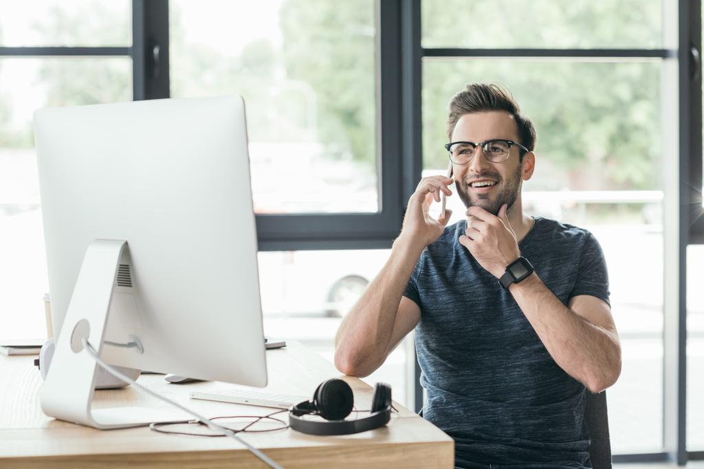 szép, mosolygós fiatalember a szemüvegek beszél a smartphone, munka közben a asztali számítógép - Fotó, kép