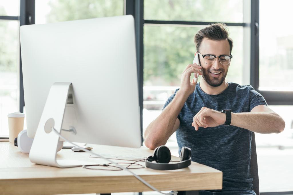 usměvavý mladý muž v brýlích kontrola smartwatch a mluví o smartphone na pracovišti - Fotografie, Obrázek