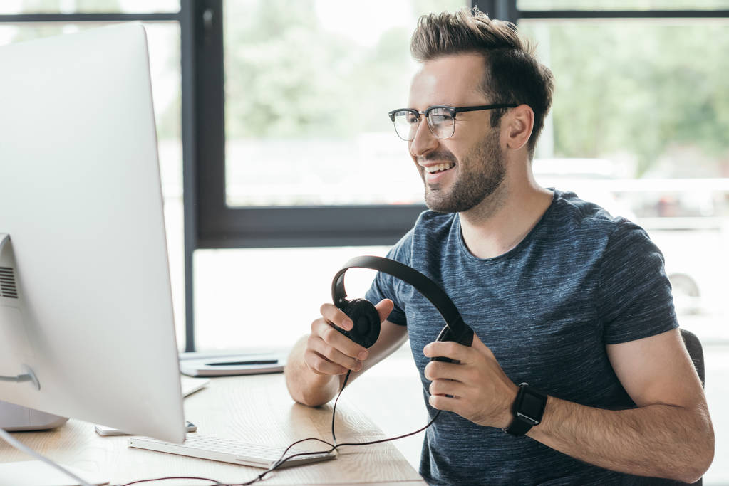 όμορφος χαμογελαστός άνθρωπος σε γυαλιά κρατώντας ακουστικά και χρησιμοποιώντας υπολογιστή - Φωτογραφία, εικόνα