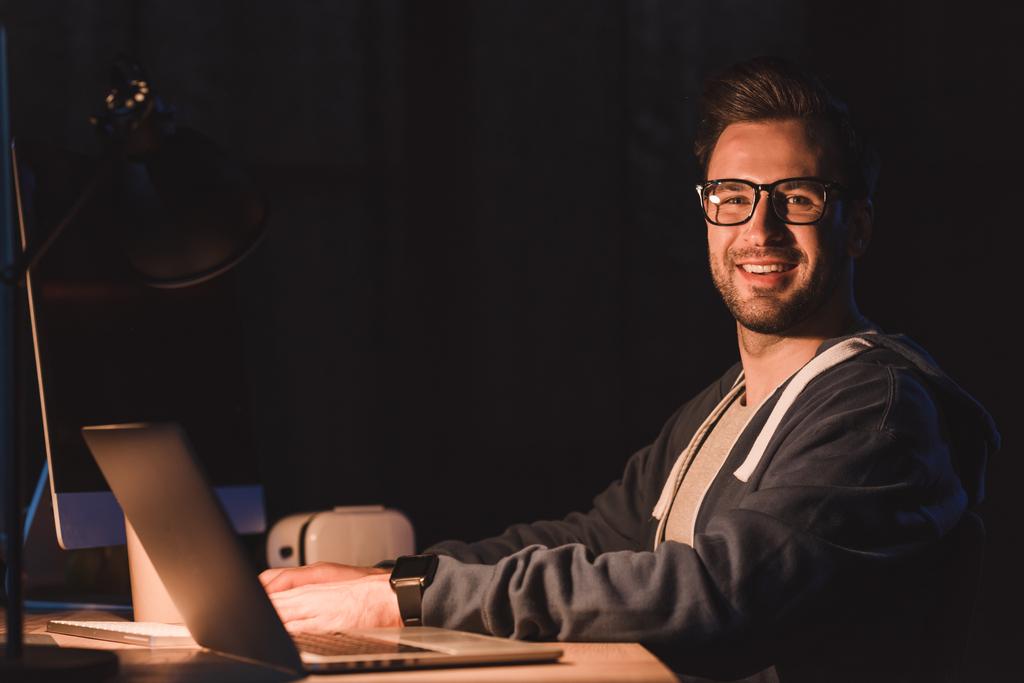pohledný mladý programátor v brýlích se usmívá na kameru při práci s počítačem laptop a desktop v noční době - Fotografie, Obrázek