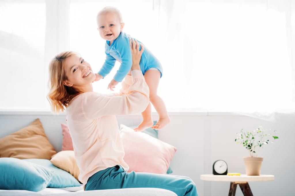 side view of smiling mother raising laughing little child while sitting on bed at home - Photo, Image