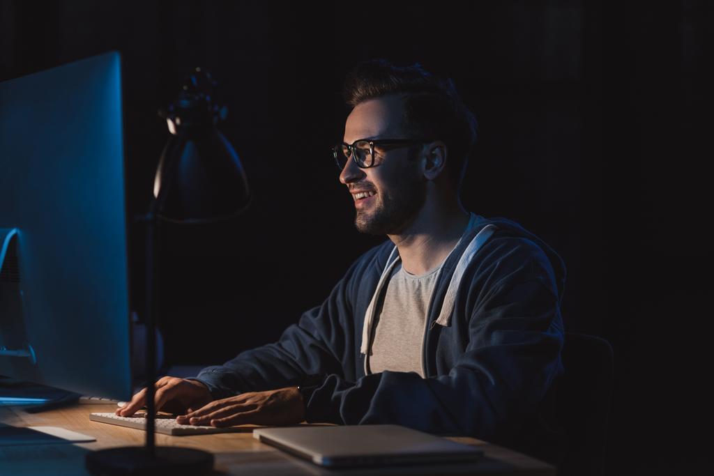 Lächelnder junger Programmierer mit Brille, der nachts am Desktop-Computer arbeitet - Foto, Bild