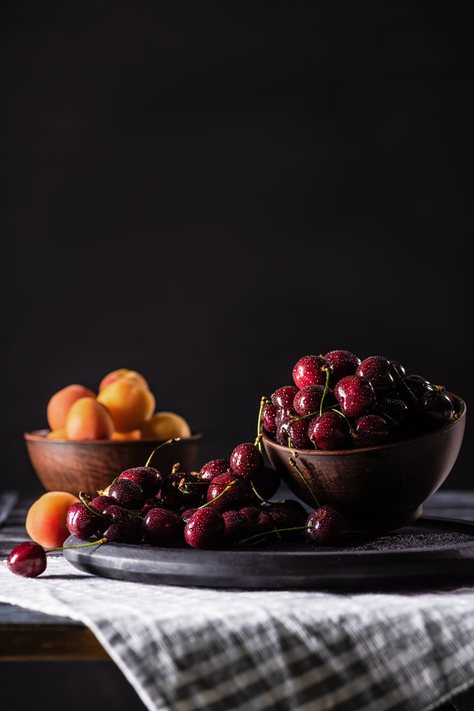 selective focus of bowls with ripe cherries and apricots on wooden table on black  - Photo, Image