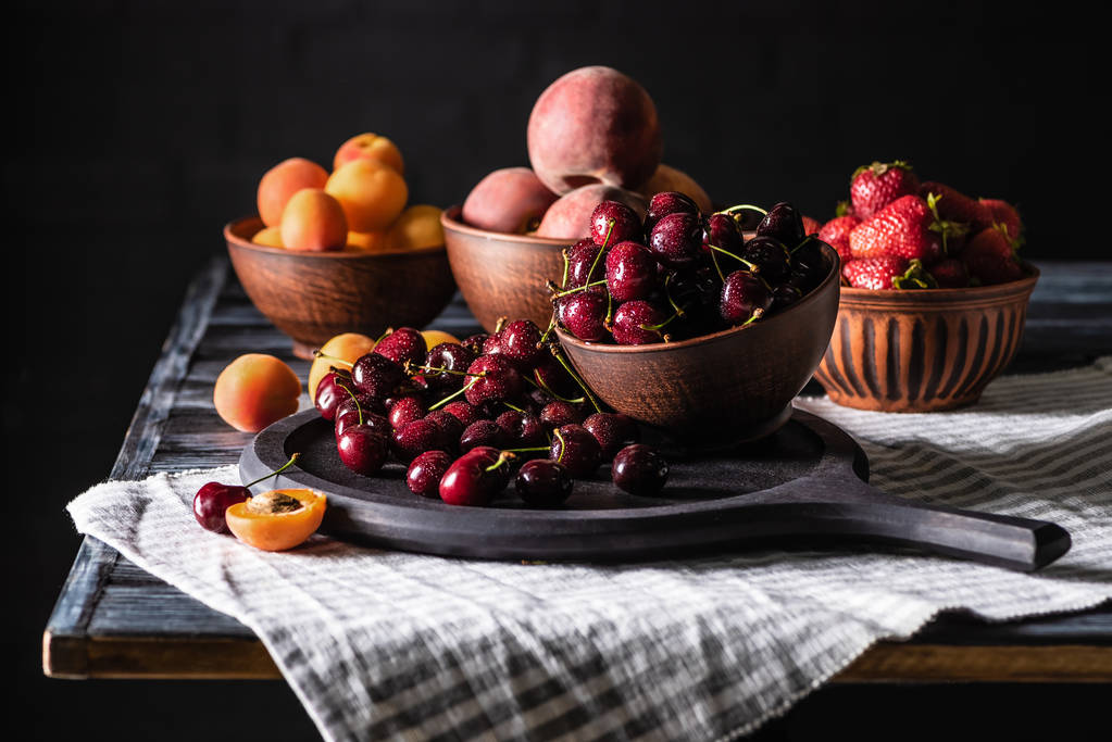 селективный фокус чаш с вишней, клубникой, персиками и абрикосами на деревянном столе на черном
  - Фото, изображение