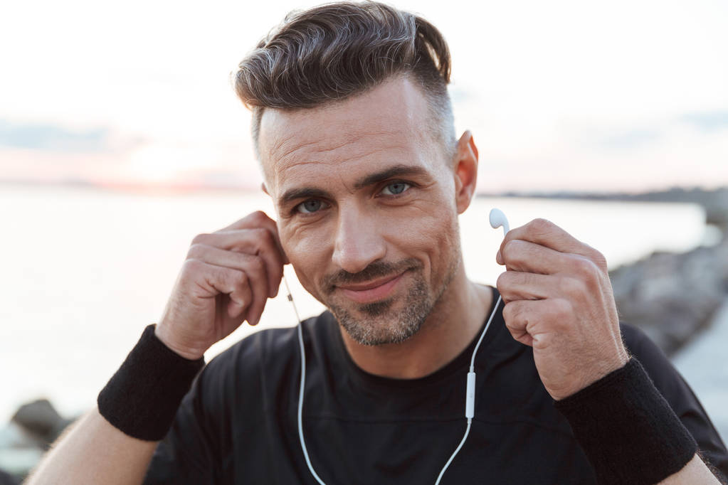 Retrato de cerca de un deportista feliz escuchando música con auriculares mientras está de pie en la playa
 - Foto, Imagen