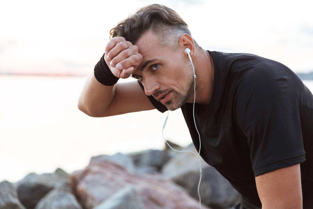 Nahaufnahme Porträt eines erschöpften Sportlers, der nach dem Joggen am Strand mit Kopfhörern Musik hört - Foto, Bild