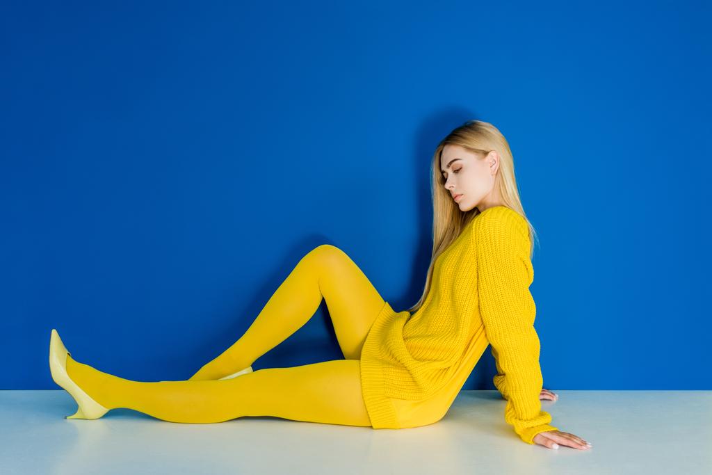 Elegant blonde woman dressed in yellow sitting on floor on blue background - Photo, Image