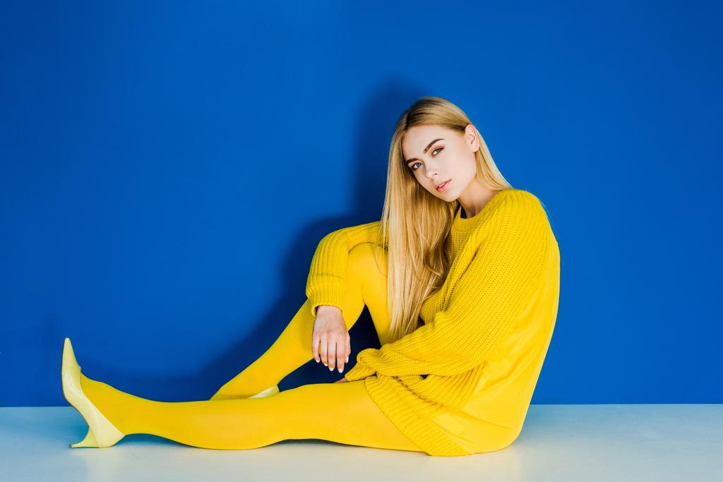 Γυναικεία μόδα μοντέλο ντυμένος στα κίτρινα κάθεται στο πάτωμα σε μπλε φόντο - Φωτογραφία, εικόνα