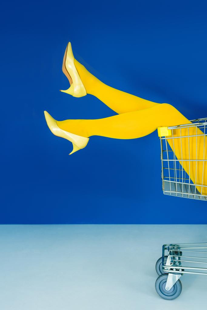 Cropped view of female legs in shopping cart on blue background - Photo, Image
