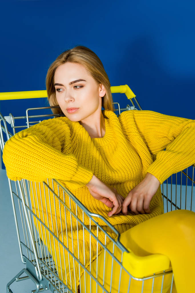 Γυναικεία μόδα μοντέλο με κίτρινα ρούχα που κάθεται στο καλάθι αγορών σε μπλε φόντο - Φωτογραφία, εικόνα