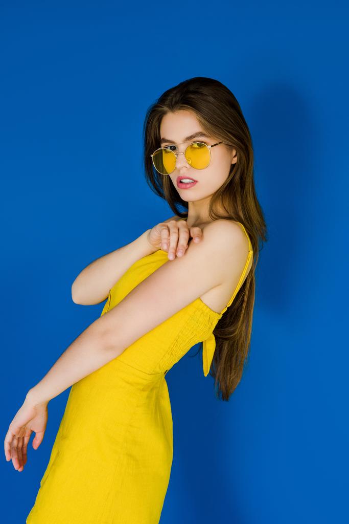 Bruneta žena ve žlutých šatech a sluneční brýle na modrém pozadí - Fotografie, Obrázek