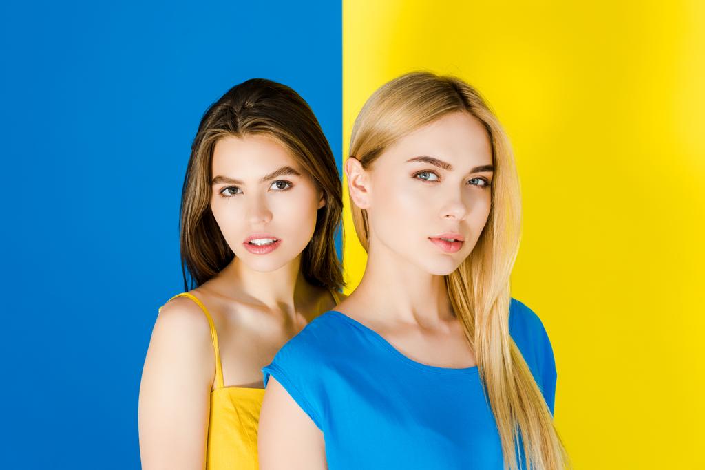 Piękna Brunetka i Blondynka dziewczyny patrząc na kamery na białym tle na tle niebieski i żółty - Zdjęcie, obraz