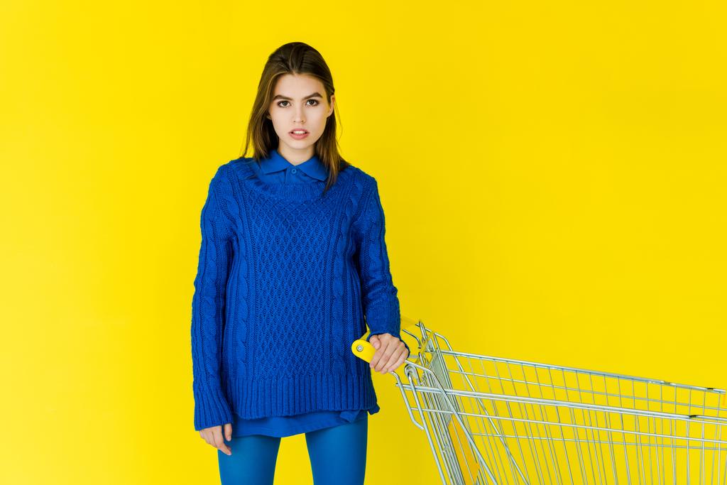 Γυναικεία μόδα μοντέλο σε μπλε πουλόβερ κρατώντας καλάθι αγορών που απομονώνονται σε κίτρινο φόντο - Φωτογραφία, εικόνα