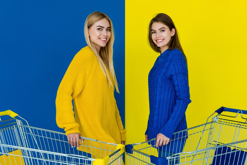 Alışveriş sepetleri ve gülümseyen mavi ve sarı zemin üzerine izole tutan çekici genç kızlar - Fotoğraf, Görsel