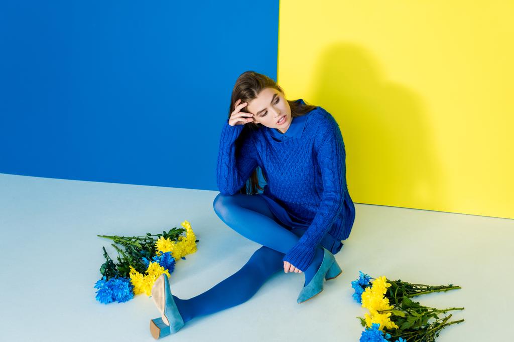 Vrouw in blauwe kleding zit onder de bloemen op de blauwe en gele achtergrond - Foto, afbeelding