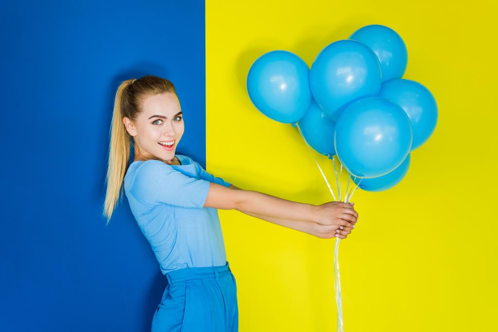 Attractive young girl holding bunch of blue balloons on blue and yellow background - Photo, Image