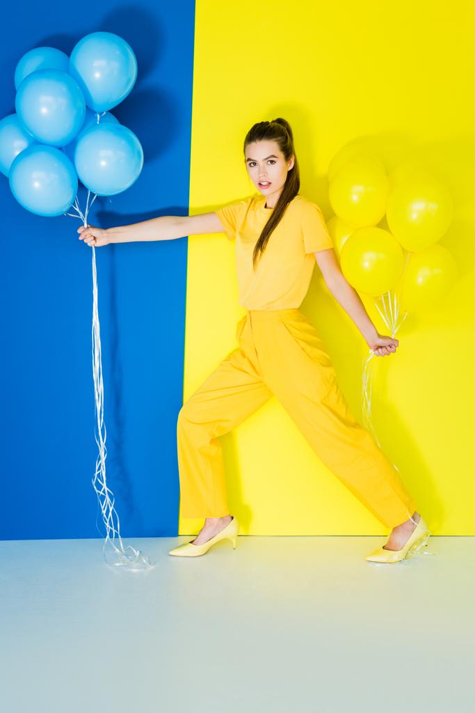 Vonzó fiatal lány gazdaság lufi, mindkét kezével a kék és a sárga háttér - Fotó, kép