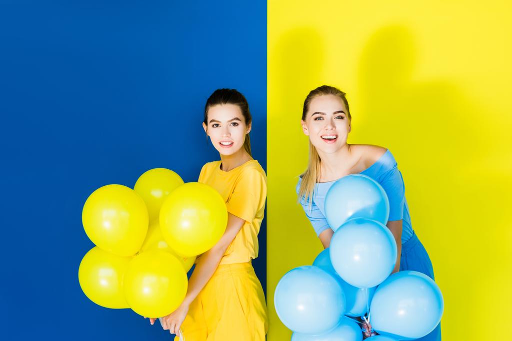 Elegante modieuze vrouwen glimlachend en houden partij ballonnen op blauwe en gele achtergrond - Foto, afbeelding