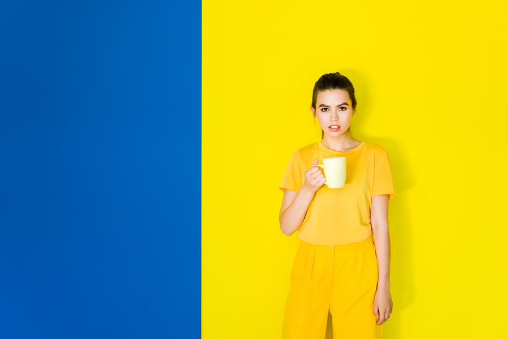 Piękna brunetka dziewczynka gospodarstwa yellow cup na tle niebieski i żółty - Zdjęcie, obraz