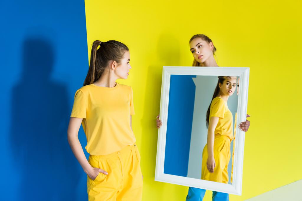Attractive young girl holding mirror for her friend on blue and yellow background - Photo, Image