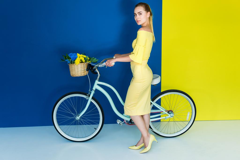 Elegáns szőke nő pózol kerékpárral, virág kosár a kék és a sárga háttér - Fotó, kép