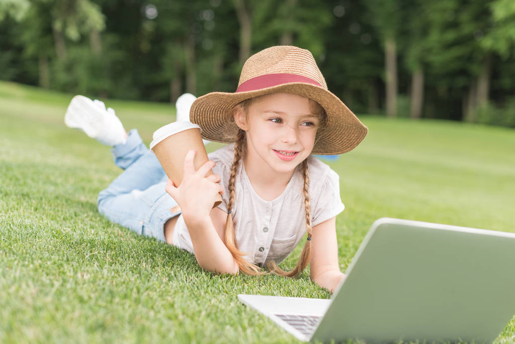 Glückliches Kind mit Pappbecher und Laptop im Park auf Gras liegend - Foto, Bild