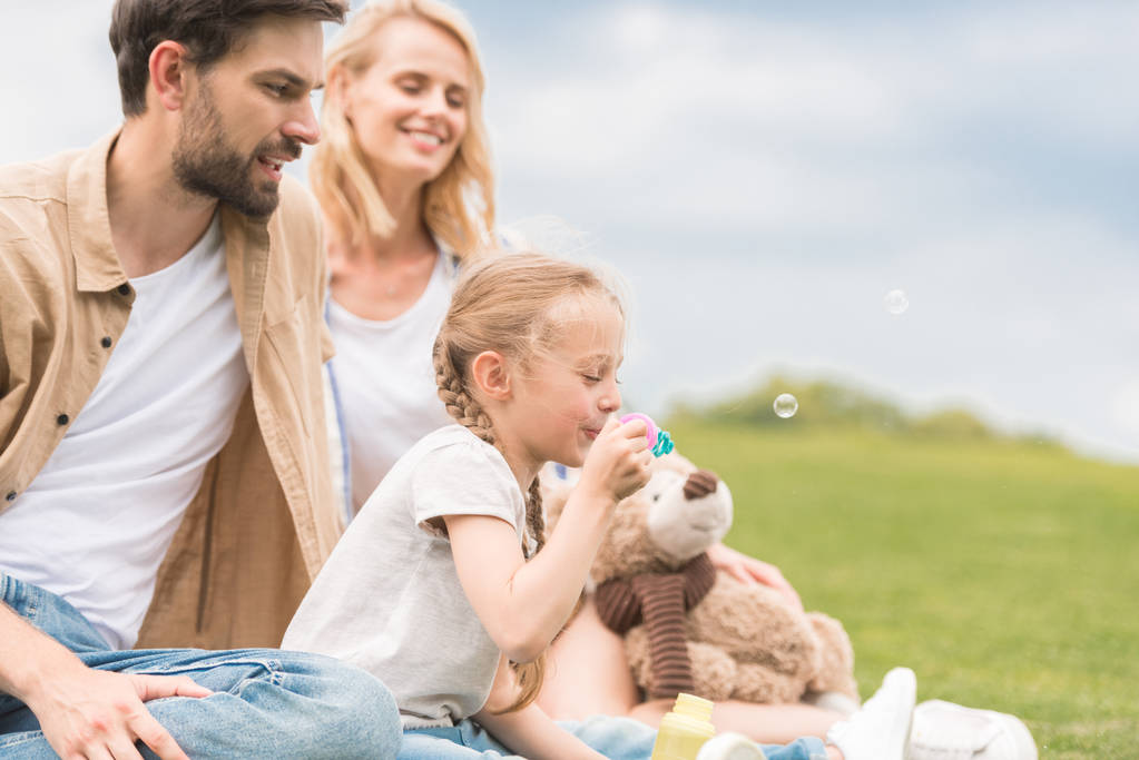 šťastní rodiče při pohledu na roztomilá malá dcerka se medvídek foukání mýdlové bubliny v parku  - Fotografie, Obrázek