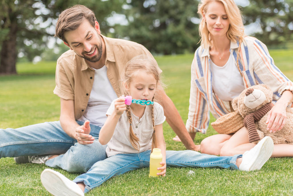 ευτυχείς γονείς βλέπουν χαριτωμένη μικρή κόρη φυσώντας φυσαλίδες σαπουνιού στο πάρκο  - Φωτογραφία, εικόνα