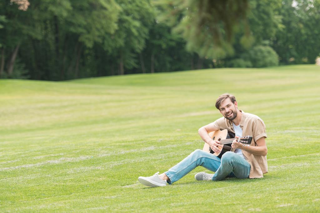 onnellinen nuori mies istuu vihreällä niityllä ja soittaa akustista kitaraa
 - Valokuva, kuva
