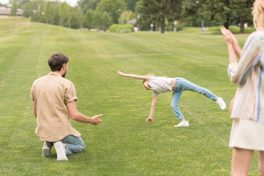 rodiče při pohledu na roztomilá malá dcera hraje na zelené louce v parku - Fotografie, Obrázek