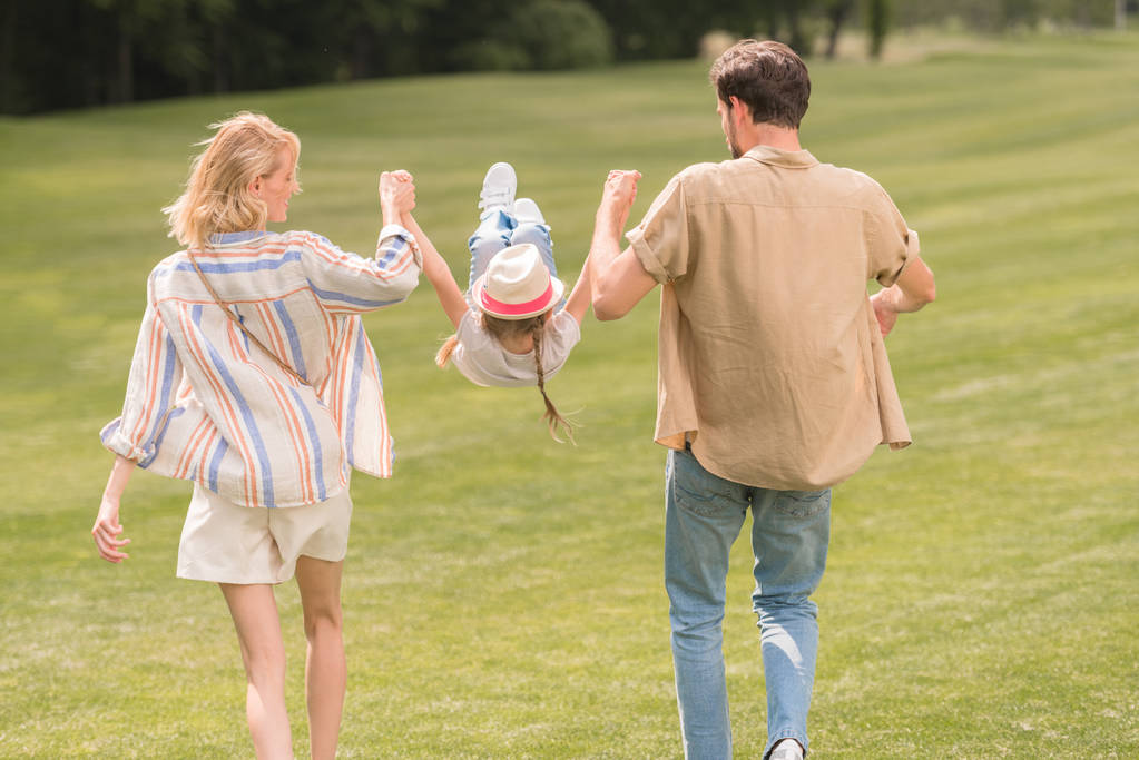 zadní pohled rodičů s malou dcerou, drželi se za ruce a baví se při chůzi spolu v parku - Fotografie, Obrázek