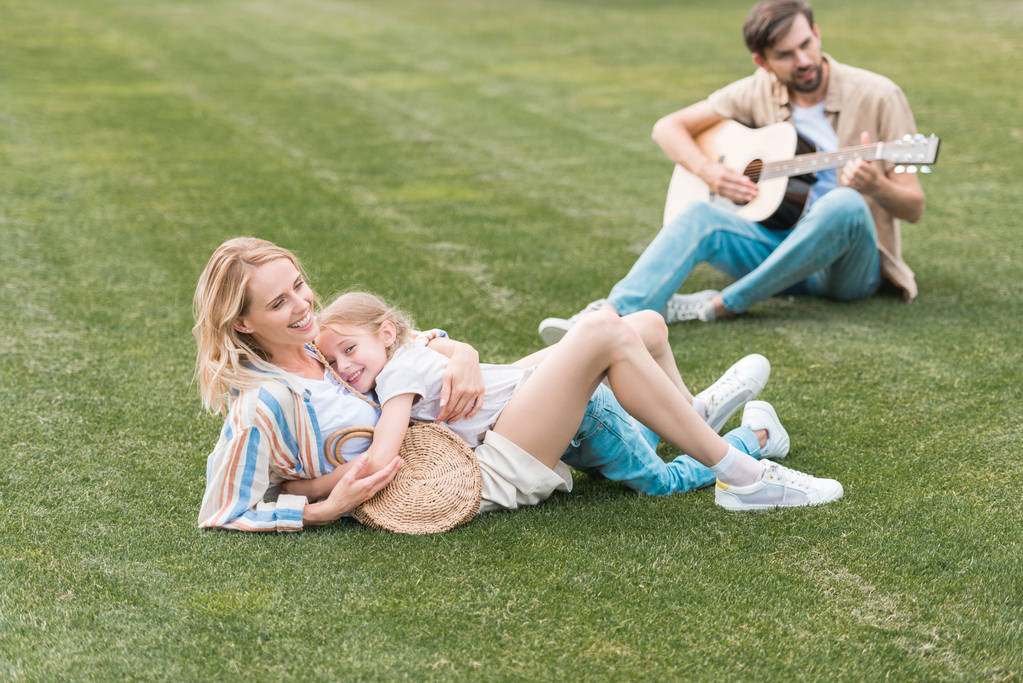 happy mother and daughter hugging on lawn while father playing guitar behind - Photo, Image