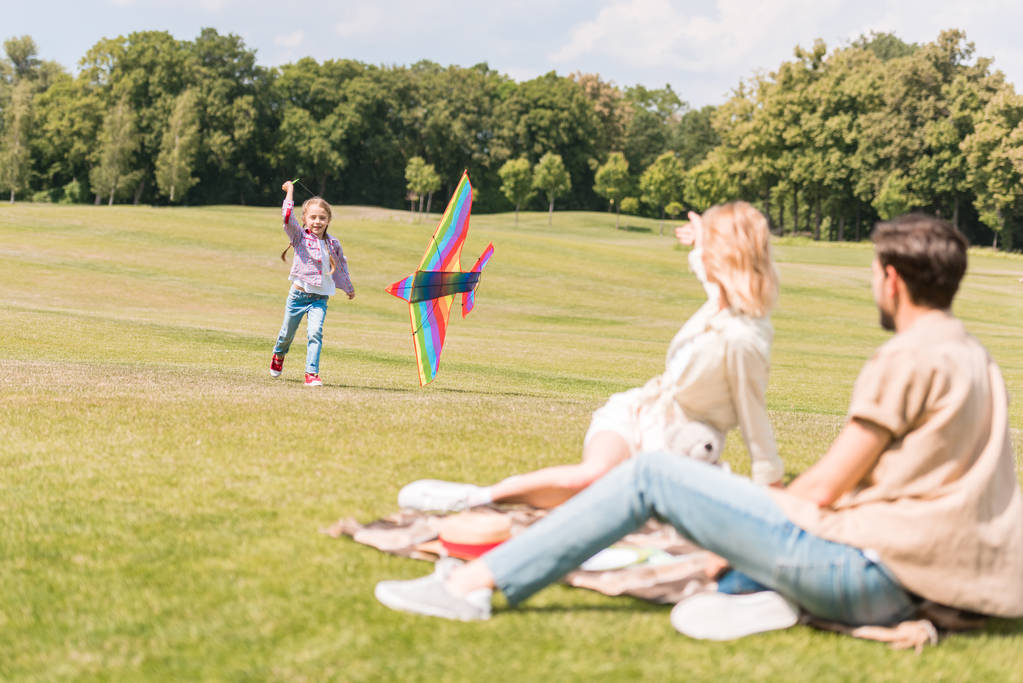 Eltern sitzen auf Plaid und schauen Tochter beim Spielen mit buntem Drachen im Park zu  - Foto, Bild