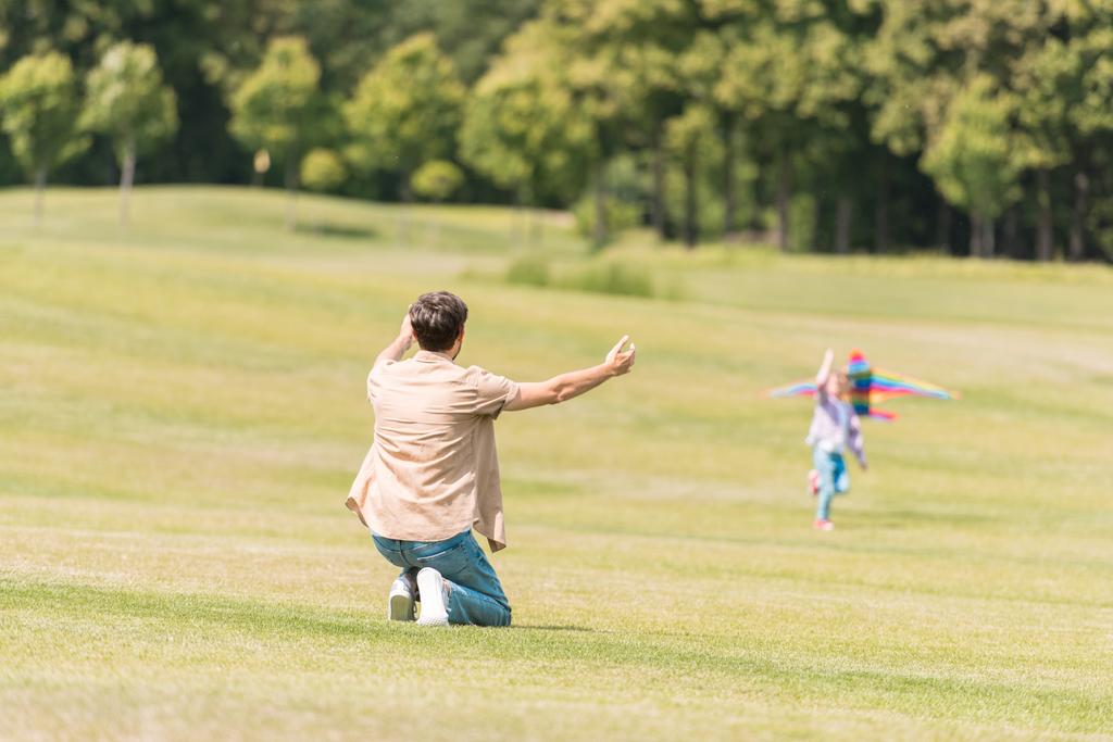 vista trasera del padre con los brazos abiertos mirando a la pequeña hija jugando con cometa en el parque
 - Foto, imagen