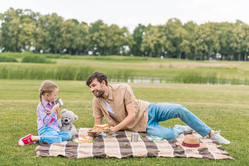 feliz padre e hija sonriendo el uno al otro mientras se sientan a cuadros en el picnic en el parque
  - Foto, Imagen