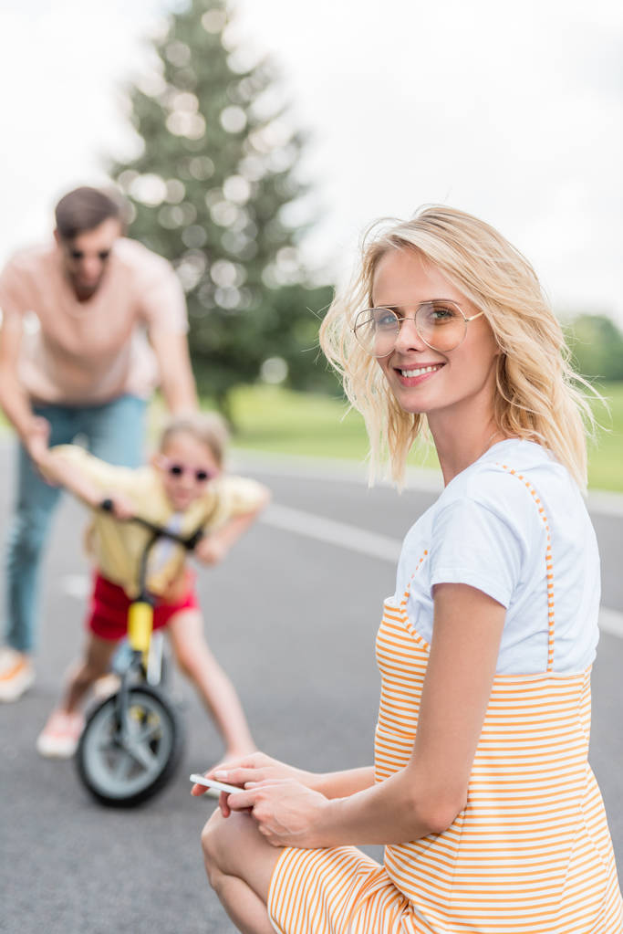Szczęśliwa młoda kobieta trzymając smartfon i uśmiecha się do kamery, podczas gdy ojciec córka nauczania jazdy rower za - Zdjęcie, obraz